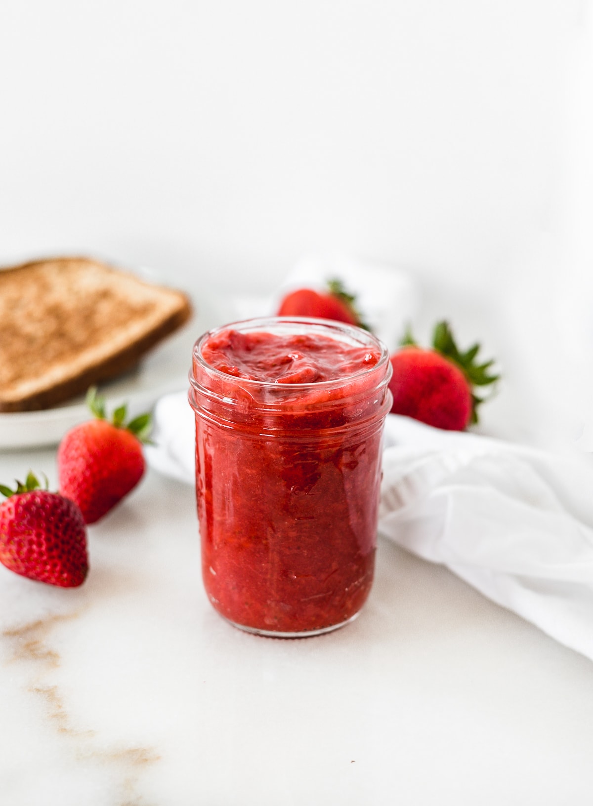 strawberry jam in a glass jar with strawberries and toast behind it.
