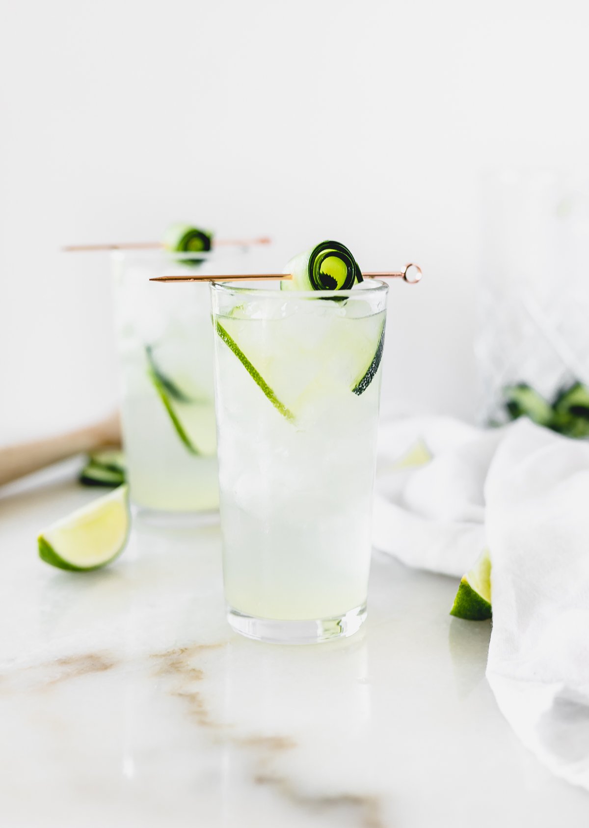 white linen cocktail in a glass with lime and cucumber slice in it topped with a rolled cucumber on a rose gold cocktail pick with another cocktail in the background.