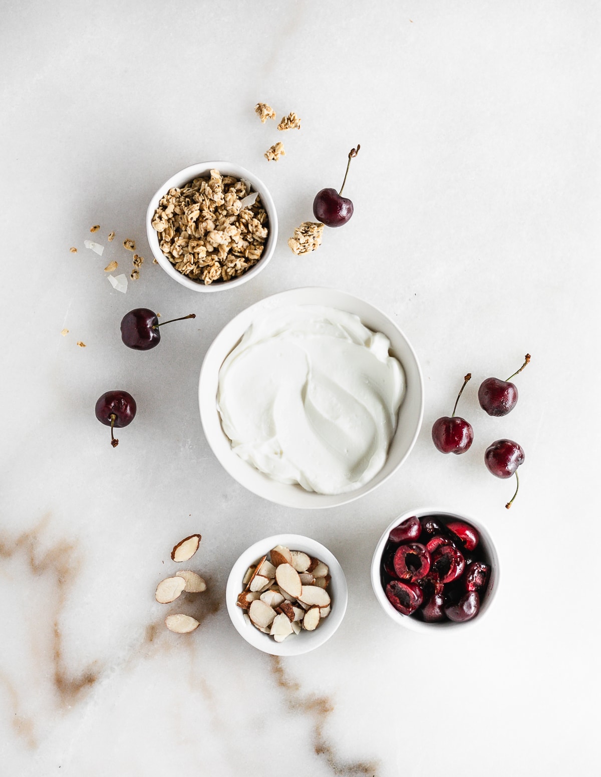 overhead view of yogurt, granola, sliced almonds, and cherries in individual bowls on a white background.