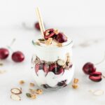 cherry almond yogurt parfait in a glass jar with a gold spoon in it.
