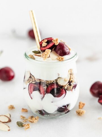 cherry almond yogurt parfait in a glass jar with a gold spoon in it.